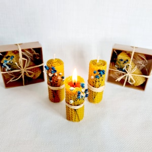 May include: Three yellow beeswax candles with dried flowers, one is lit, all are in a honeycomb pattern, two are in brown boxes with twine and a bow