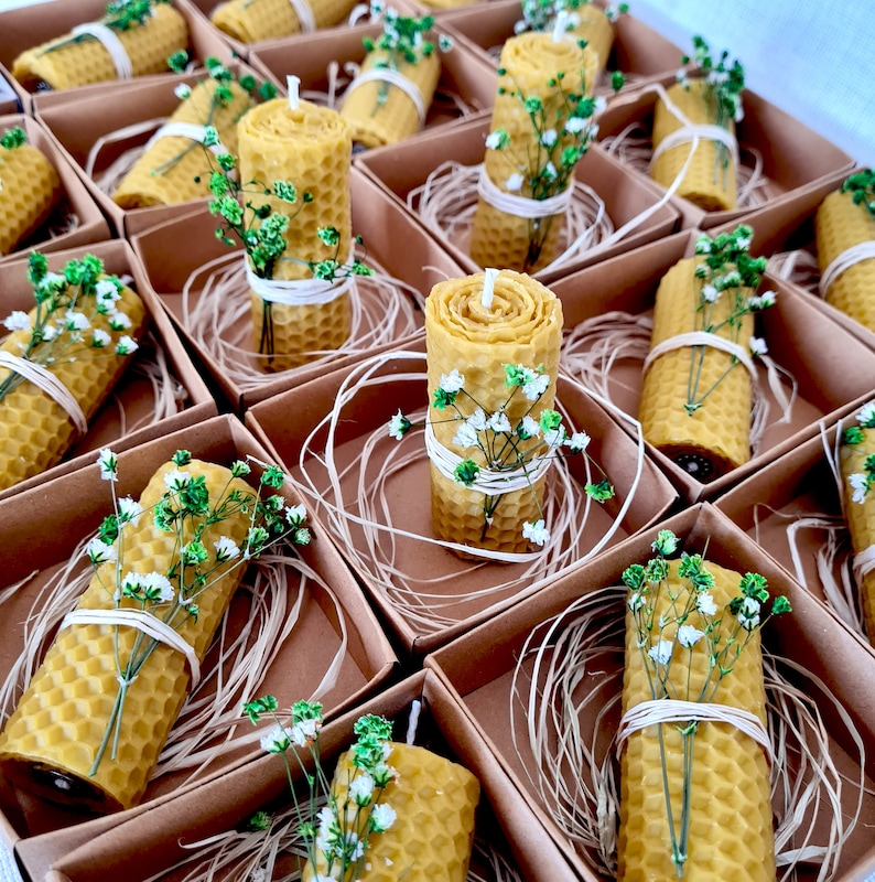 May include: A collection of small, natural beeswax candles in individual boxes. Each candle is decorated with a sprig of white flowers and wrapped in twine. The boxes are lined with straw-like material.
