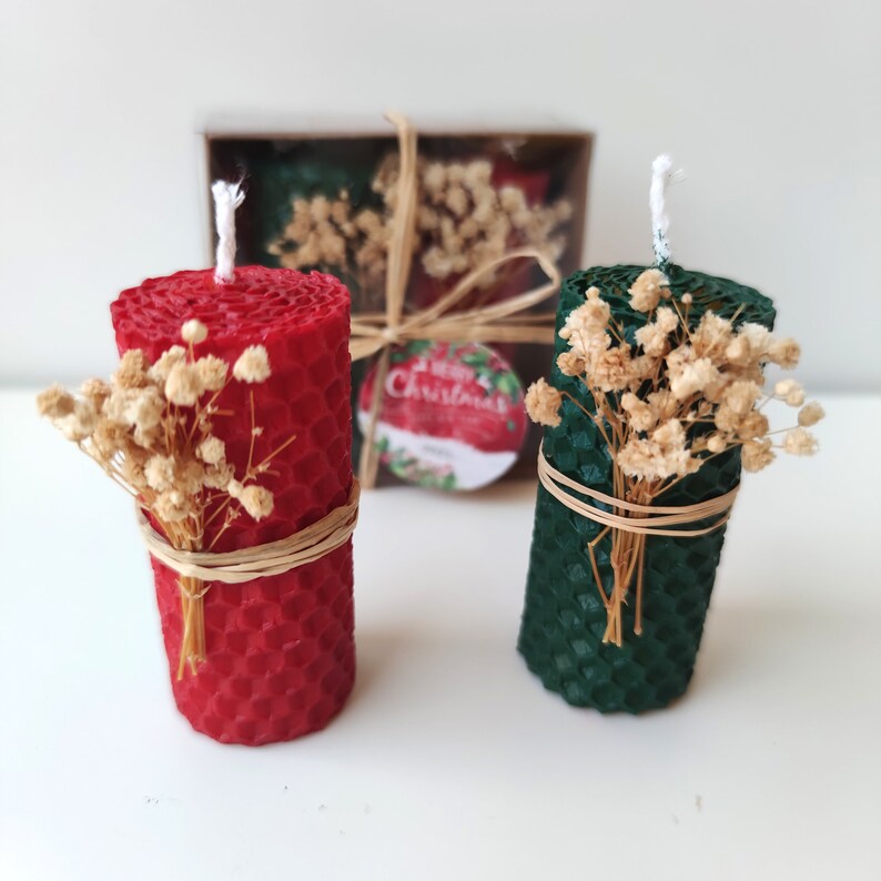 May include: Two small honeycomb candles, one red and one green, decorated with dried flowers and tied with twine. The candles are in a box with a "Merry Christmas" label.