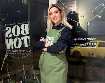 May include: An olive green apron featuring the white text BOS TON on the chest. The apron has a tie waist and is worn over a black hooded sweatshirt, black tights, and black lace-up boots with brown laces and a side logo detail.