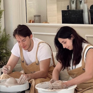Puede incluir: Dos personas trabajando en tornos de alfarero, dando forma a la arcilla. Llevan camisetas blancas y delantales color beige. La escena se desarrolla en un estudio luminoso con estanterías de cerámica y objetos decorativos. Los delantales tienen la palabra "US".