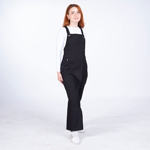 Puede incluir: Un vestido de peto negro con tirantes ajustables y un bolsillo delantero. El vestido tiene un corte holgado y una pierna ancha.