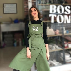 Könnte beinhalten: Eine Frau trägt eine grüne Schürze mit dem Schriftzug "BOSTON" in weißen Buchstaben. Sie steht vor einem Ladenfenster mit dem Wort "BOSTON" darauf.