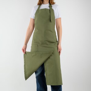 Könnte beinhalten: Olivgrüne Schürze mit Wickeldesign und großer Fronttasche. Die Schürze hat einen Nackenriemen und einen Taillenverschluss. Das Model trägt ein weißes T-Shirt und blaue Jeans. Die Schürze ist aus strapazierfähigem Stoff gefertigt.