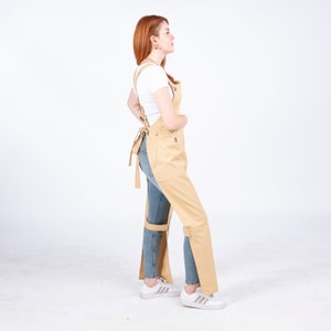 Puede incluir: Un delantal de lona beige con tirantes ajustables y una correa para la pierna. El delantal se lleva sobre unos vaqueros azules y una camiseta blanca.