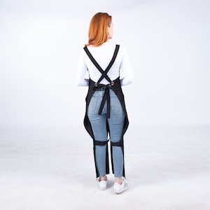 Puede incluir: Delantal negro con correas cruzadas y correas para las piernas. El delantal se usa sobre una camisa blanca de manga larga y jeans azules. El delantal tiene una cinturilla y ojales metálicos.