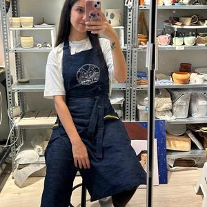 Puede incluir: Un delantal de mezclilla azul oscuro con un logotipo circular, usado sobre una camiseta blanca. El delantal tiene detalles amarillos y un lazo en la cintura. Estanterías en el fondo exhiben cerámica y artículos de cerámica.