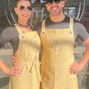 Puede incluir: Dos personas con delantales amarillos claros a juego con el texto "DESI at house" impreso. Los delantales tienen bolsillos y están atados a la cintura. Las personas sonríen y llevan gafas de sol.