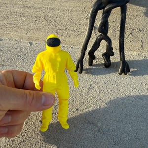 Op de afbeelding: Een kleine, gele hazmat-pak figuur met een zwart vizier wordt in een hand gehouden. Een zwarte, abstracte sculptuur met lange, gedraaide ledematen staat in de buurt. De figuren staan tegen een getextureerde, lichtgekleurde achtergrond.