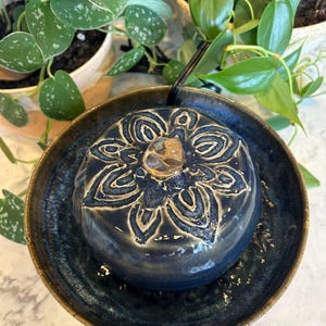 May include: A dark blue ceramic water fountain featuring a raised, carved floral pattern on the top dome. The fountain sits in a matching circular basin with a glossy, textured glaze. A clear, rounded stone sits at the centre of the floral design.
