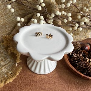 May include: A pair of gold-toned stud earrings featuring the text FUCK OFF in a bold, sans-serif font. The earrings are displayed on a white, scalloped-edge ceramic pedestal stand, positioned centrally on the smooth, flat surface of the stand.
