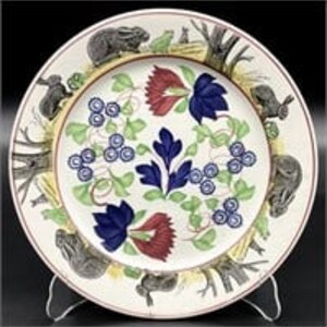 May include: Decorative ceramic plate with a central floral design in blue, red, and green. The rim features illustrations of animals and trees. The plate is displayed on a clear stand.