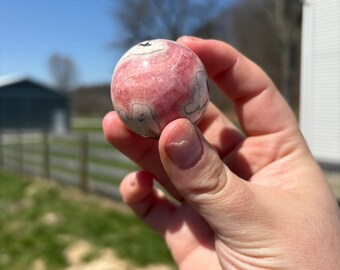 May include: A pink and white sphere-shaped gemstone, held in a hand, against a backdrop of a sunny day. The stone has a swirling pattern of pink and white. The background includes a green field, a fence, and a building.