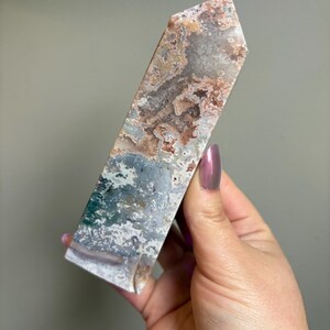 May include: A polished, rectangular crystal point with a flat top. The stone features a mix of colors, including brown, white, green, and gray, with a marbled appearance. The crystal is held in a hand.