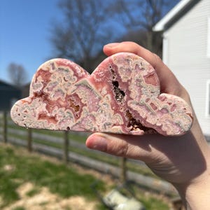 May include: A cloud-shaped decorative stone with pink, white, and brown patterns. The stone is held up in front of a sunny, outdoor background. The stone has a rough, natural texture and a unique shape.