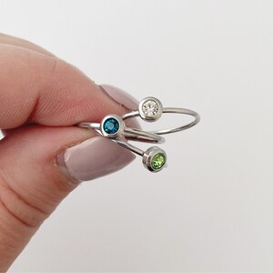 May include: Three silver rings with small colorful gemstones. The rings are stacked on top of each other. The top ring has a clear gemstone, the middle ring has a blue gemstone, and the bottom ring has a green gemstone.