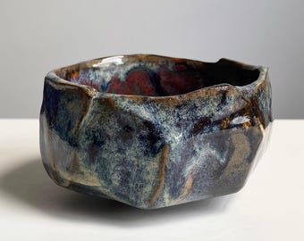 May include: A handmade ceramic bowl with a unique, faceted design. The bowl features a blend of blue, brown, and red glaze, creating a textured, organic appearance. The interior has a deep red hue, contrasting with the exterior's cooler tones. A decorative piece for the home.