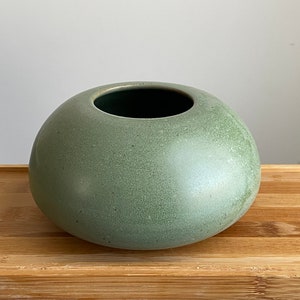 May include: A round, green ceramic vase with a speckled finish. The vase has a wide opening and a smooth, rounded bottom.