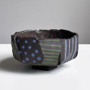 May include: A dark, geometric ceramic bowl with a square base. The bowl's exterior features sections with blue polka dots, green horizontal stripes, and blue vertical stripes. The interior has green polka dots.