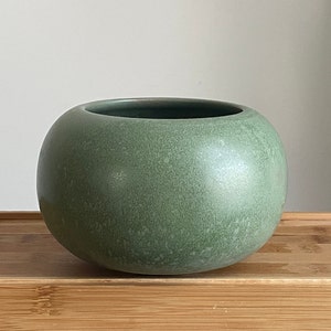 May include: A green ceramic planter with a speckled glaze. The planter is round and has a wide opening.