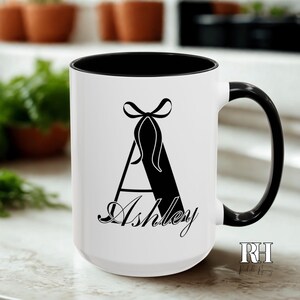 Pode incluir: Caneca de cerâmica branca com interior e alça pretos. A caneca apresenta a letra "A" preta com um desenho de laço e o nome "Ashley" em fonte cursiva. A caneca tem um tamanho padrão para café ou chá.