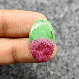 Pode incluir: Uma pedra preciosa oval, dividida em duas cores distintas. A metade superior é verde brilhante, a inferior vermelha intensa. A pedra polida é segurada por um dedo e tem um acabamento brilhante.
