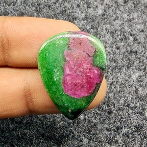Pode incluir: Uma pedra preciosa polida com formato de gota. A pedra apresenta uma borda verde vibrante em torno de uma área central com tons rosa e vermelho profundos. A pedra é segurada em uma mão.