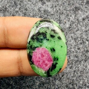 Pode incluir: Gema oval com um centro rosa vibrante rodeado por padrões verdes e pretos salpicados. A pedra polida é segurada em uma mão, mostrando sua beleza natural e combinação de cores única.