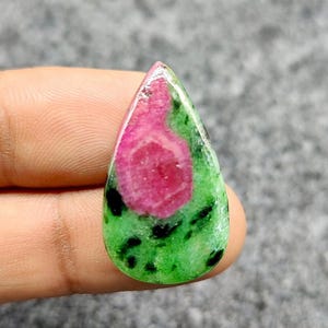 Pode incluir: Uma pedra preciosa em forma de gota, com um centro rosa vibrante e um exterior verde salpicado de pontos pretos. A pedra polida é segurada, mostrando suas cores e padrões naturais. Esta gema única é ideal para fazer joias.
