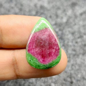 Pode incluir: Uma pedra preciosa em forma de gota com uma paleta de cores vibrantes. A pedra apresenta uma área central de rosa intenso, circundada por uma faixa de verde brilhante e uma camada externa translúcida. A superfície polida reflete a luz.