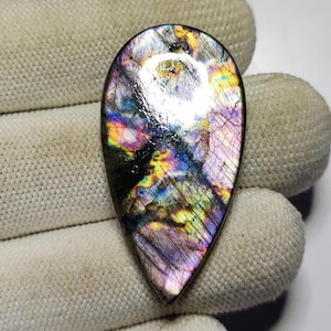 May include: A polished, teardrop-shaped labradorite gemstone. The stone exhibits a shimmering play of colour, including blues, purples, greens, and yellows. The gemstone is set against a white background.
