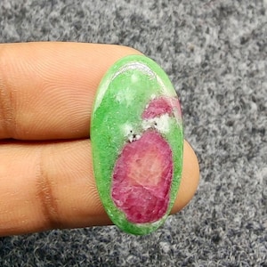 Pode incluir: Gema ovalada com uma base verde vibrante e uma inclusão de rubi proeminente e irregular. O rubi exibe um tom vermelho profundo, contrastando com o verde mais claro. A pedra é segurada entre os dedos.