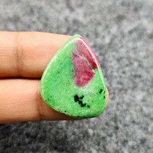 Pode incluir: Uma pedra preciosa polida em forma de gota. A pedra apresenta uma vibrante cor de base verde com uma proeminente mancha de vermelho profundo e pequenas inclusões pretas. A pedra preciosa é segurada em uma mão.