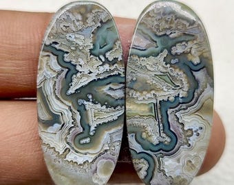 May include: Two oval-shaped gemstones with intricate patterns. The stones feature a mix of colors, including shades of gray, white, green, and tan, creating a marbled effect. The polished surfaces showcase the natural formations within the stones.