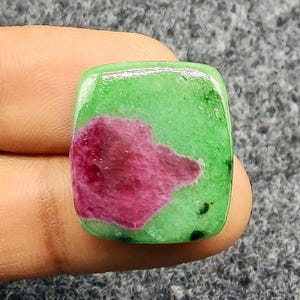 Pode incluir: Uma pedra preciosa de corte quadrado com uma base verde vibrante e um padrão magenta-vermelho proeminente e irregular. A pedra tem uma superfície polida e lisa, e as cores são ricas e saturadas. A pedra preciosa é segurada entre os dedos.