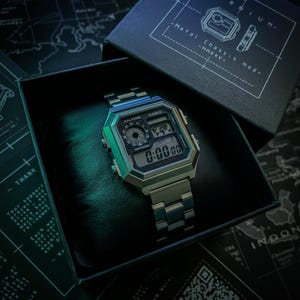 May include: A silver Casio digital watch with a metal strap is presented in a black box. The watch face shows the time. The box is labelled "-KARNIUM-" and "Metal Chassis mod-MMXXV-". The watch rests on a map background.
