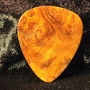 Op de afbeelding: Een gitaar plectrum met een uniek houtnerfpatroon in amber- en bruintinten. De plectrum heeft een glad, gepolijst oppervlak en een druppelvorm. De achtergrond is een donkere, getextureerde stof.