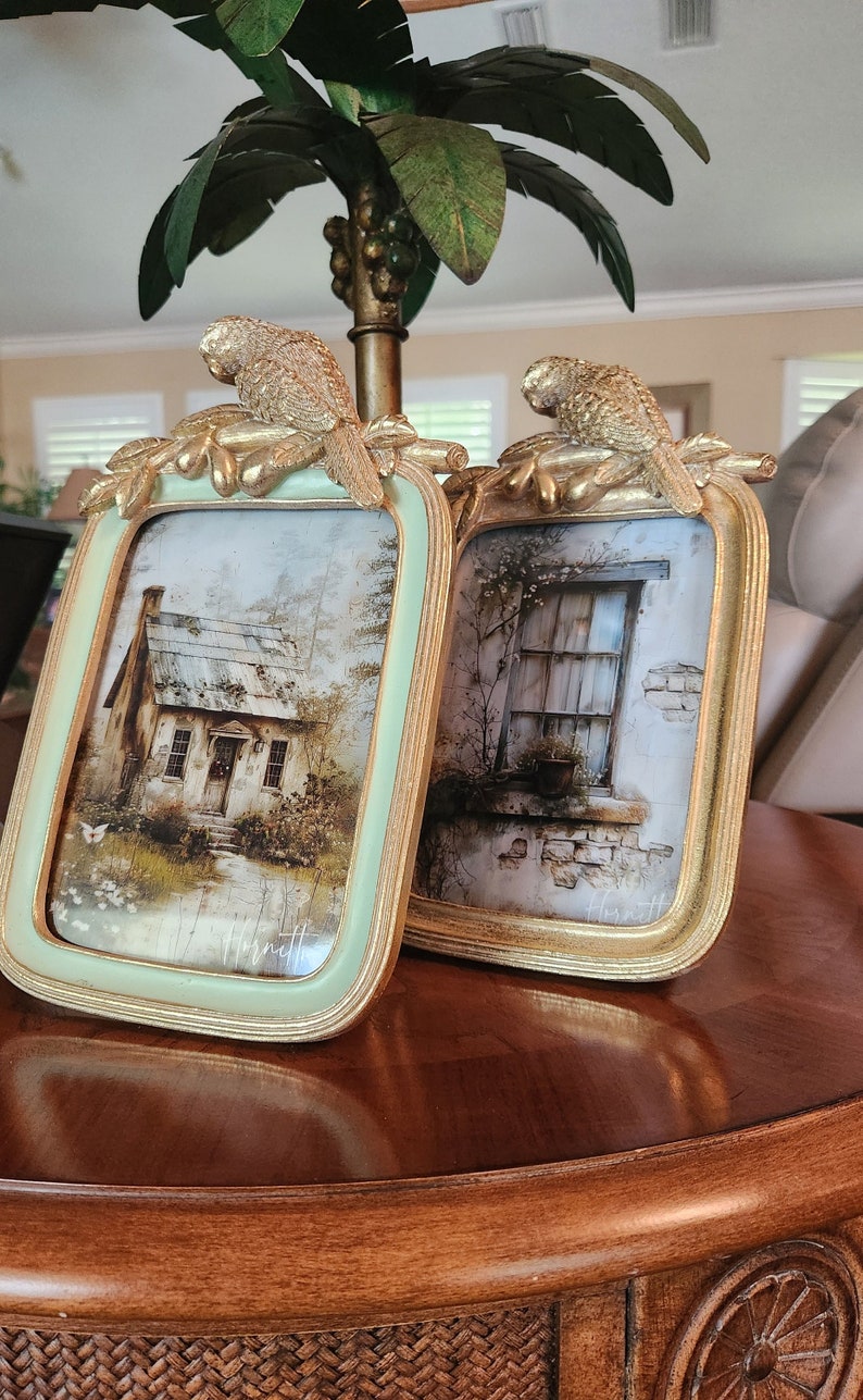 May include: Two ornate gold picture frames with bird accents and a light green background. The frames are on a wooden table. One frame features a watercolor painting of a small cottage, the other a window with a potted plant.