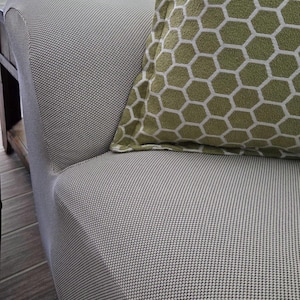 May include: Close-up of a light gray upholstered sofa with a textured pattern. A green pillow with a white honeycomb pattern rests on the sofa. The sofa is in a well-lit room.