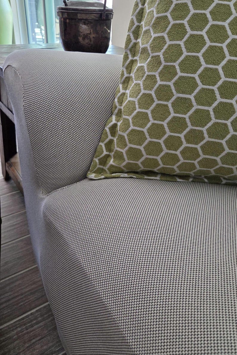 May include: Close-up of a light gray upholstered sofa with a textured pattern. A green pillow with a white honeycomb pattern rests on the sofa. The sofa is in a well-lit room.