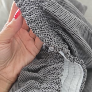 May include: Close-up of gray fabric with a textured, dotted pattern. The fabric is gathered at the edge, revealing a white elastic band. The material appears soft and pliable, suitable for clothing or home textiles.