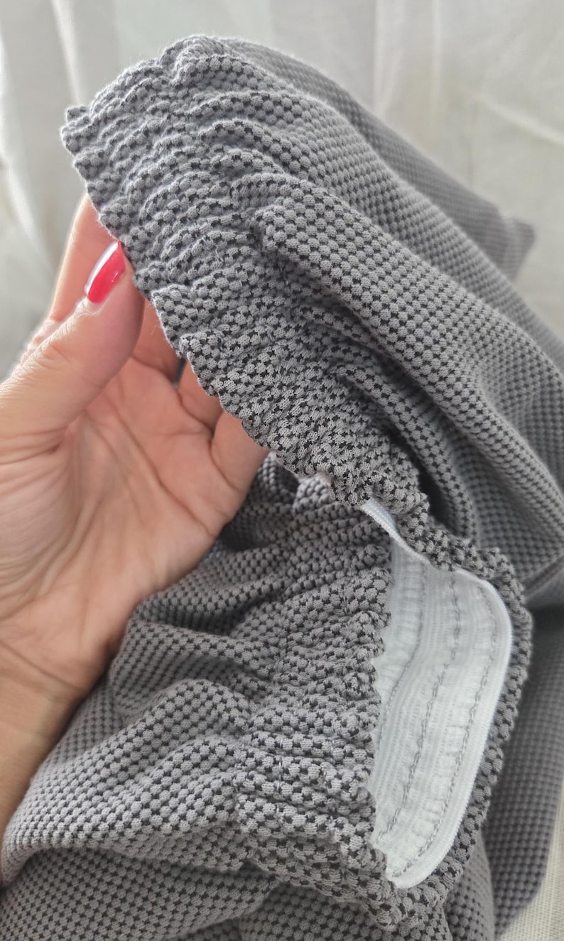 May include: Close-up of gray fabric with a textured, dotted pattern. The fabric is gathered at the edge, revealing a white elastic band. The material appears soft and pliable, suitable for clothing or home textiles.
