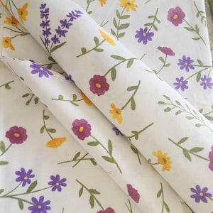 Könnte beinhalten: Weißer Stoff mit einem Blumenmuster mit violetten, gelben und rosa Blüten. Der Stoff ist in der Mitte gefaltet, sodass das Muster auf beiden Seiten sichtbar ist.