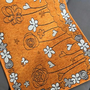 May include: An orange floral patterned rug with white, gray, and black accents. The rug features a repeating pattern of flowers, hearts, and abstract shapes. The border is lined with white and gray flowers.