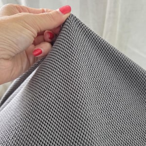 May include: Close-up of a gray fabric with a textured, dotted pattern. The fabric is held by a hand with red nail polish. The fabric appears to be soft and flexible, with a subtle sheen.