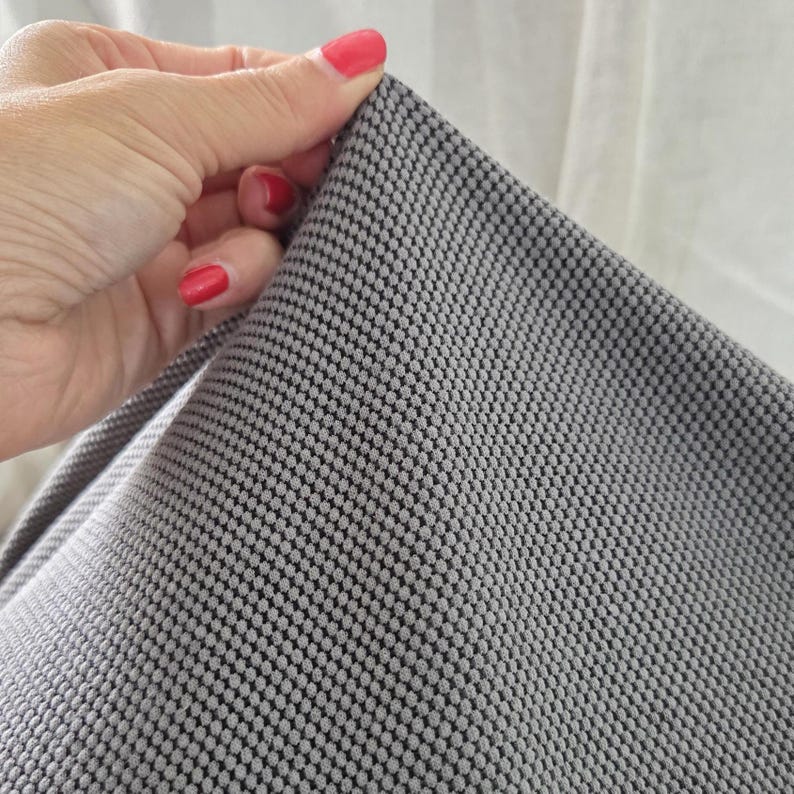 May include: Close-up of a gray fabric with a textured, dotted pattern. The fabric is held by a hand with red nail polish. The fabric appears to be soft and flexible, with a subtle sheen.