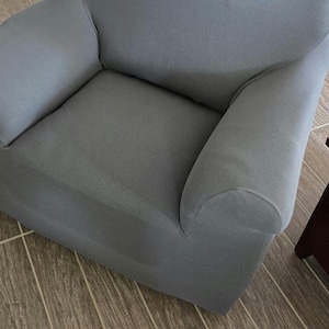 May include: A gray armchair with rounded armrests. The chair is covered in a solid gray fabric and sits on a wooden floor. A small wooden side table is partially visible in the frame.