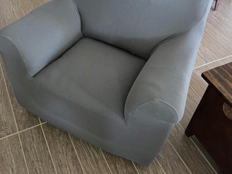 May include: A gray armchair with rounded armrests. The chair is covered in a solid gray fabric and sits on a wooden floor. A small wooden side table is partially visible in the frame.