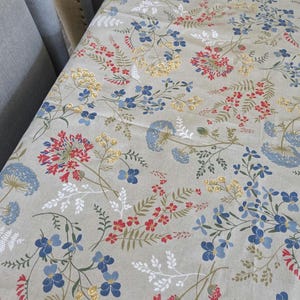 May include: A beige floral tablecloth featuring a repeating pattern of red, blue, and yellow flowers with green leaves. The flowers are of varying sizes and shapes, creating a natural, botanical design. The tablecloth is made of a soft fabric.