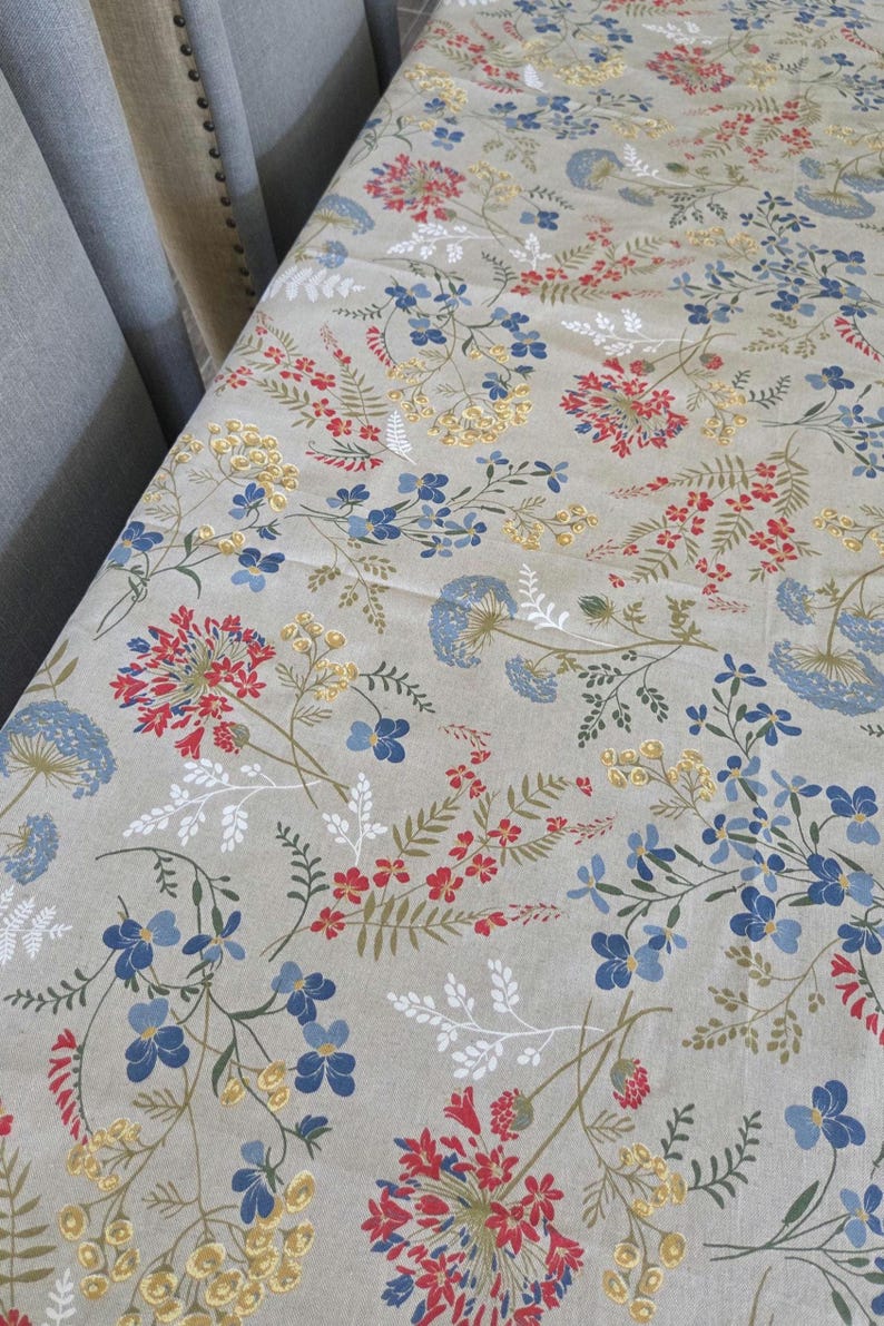 May include: A beige floral tablecloth featuring a repeating pattern of red, blue, and yellow flowers with green leaves. The flowers are of varying sizes and shapes, creating a natural, botanical design. The tablecloth is made of a soft fabric.